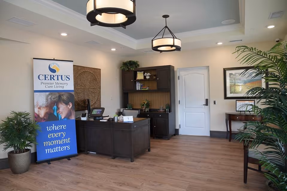 Reception area of CERTUS Premier Memory Care Living with a dark wood desk and matching cabinet, a white door, potted plants, framed artwork on the wall, and a standing banner displaying the facility's name and slogan 'where every moment matters'.