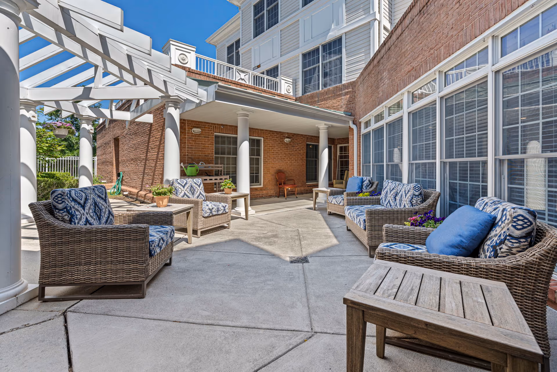 Sunlit outdoor courtyard patio with wicker seating and blue patterned cushions under a pergola beside a brick building with large windows.