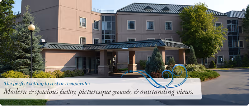 Exterior view of a modern, multi-story rehabilitation and nursing facility with beige walls, green metal roofing, surrounded by landscaped greenery including trees and shrubs under a clear sky.