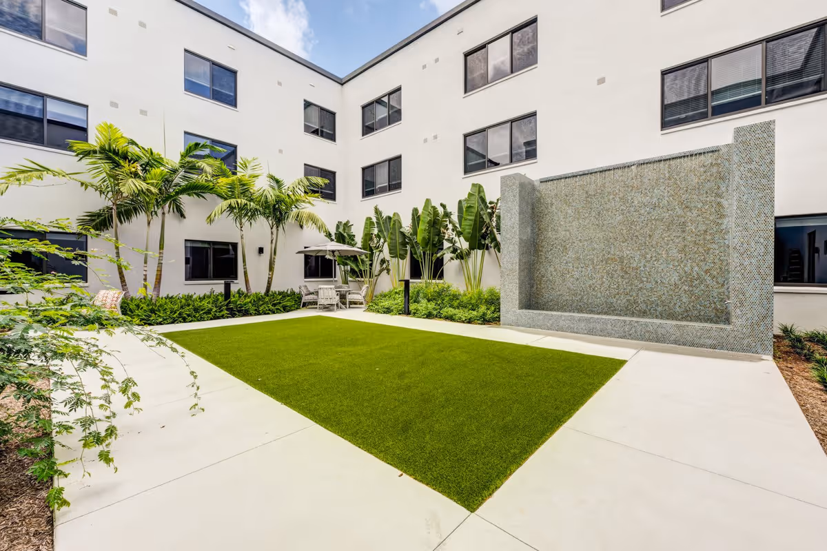 Outdoor courtyard area at The Arbor at Delray featuring a rectangular patch of green grass surrounded by concrete walkways, tropical plants including palm trees, a tiled water feature wall, and a seating area with chairs and an umbrella.