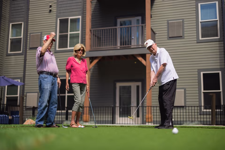 Three elderly people playing mini golf on a putting green outside a residential building. One man is putting while the other man and woman watch. The building has gray siding, multiple windows, and a balcony.