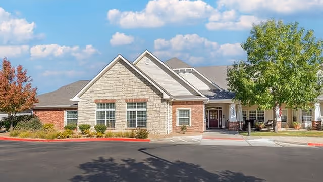 Exterior front view of a single-story senior living facility building with a combination of stone and brick facade, large windows, a covered entrance, and surrounding landscaping including trees and shrubs under a partly cloudy sky.
