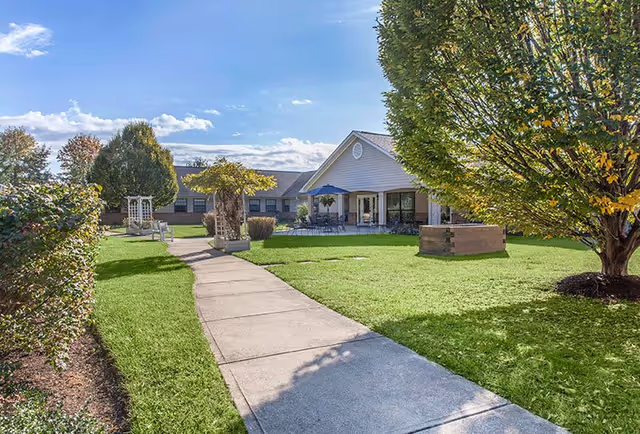 A sunny outdoor garden area at Charter Senior Living of Columbia featuring a concrete walkway leading to a building with a covered patio. The patio has tables with umbrellas and chairs. The garden includes green grass, trees, bushes, and a wooden planter box.