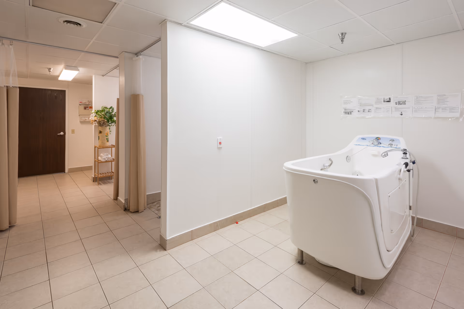 A clean, tiled room with a large white walk-in bathtub designed for assisted bathing. The room has white walls and a drop ceiling with fluorescent lighting. To the left, there is a hallway with a dark wooden door and a small shelf holding a plant and some towels. The space appears to be part of a nursing or rehabilitation center.