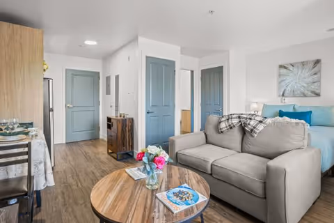 Bright studio apartment interior with a gray sofa, round wooden coffee table topped with flowers and magazines, a bed in the background, dining area, and blue doors.