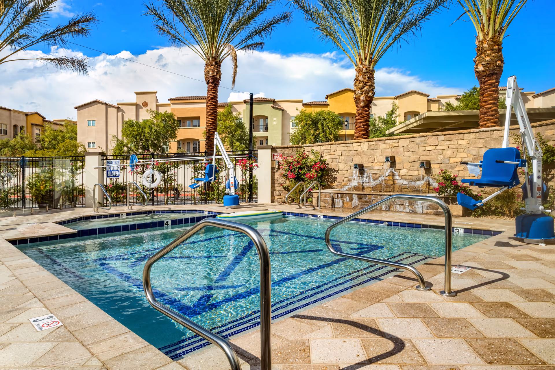 Outdoor swimming pool area with clear water, surrounded by a tiled deck and metal handrails. Two blue pool lifts for accessibility are positioned on opposite sides of the pool. Palm trees and flowering bushes are planted around the pool area, with a stone wall and multi-story residential buildings in the background under a blue sky with some clouds.