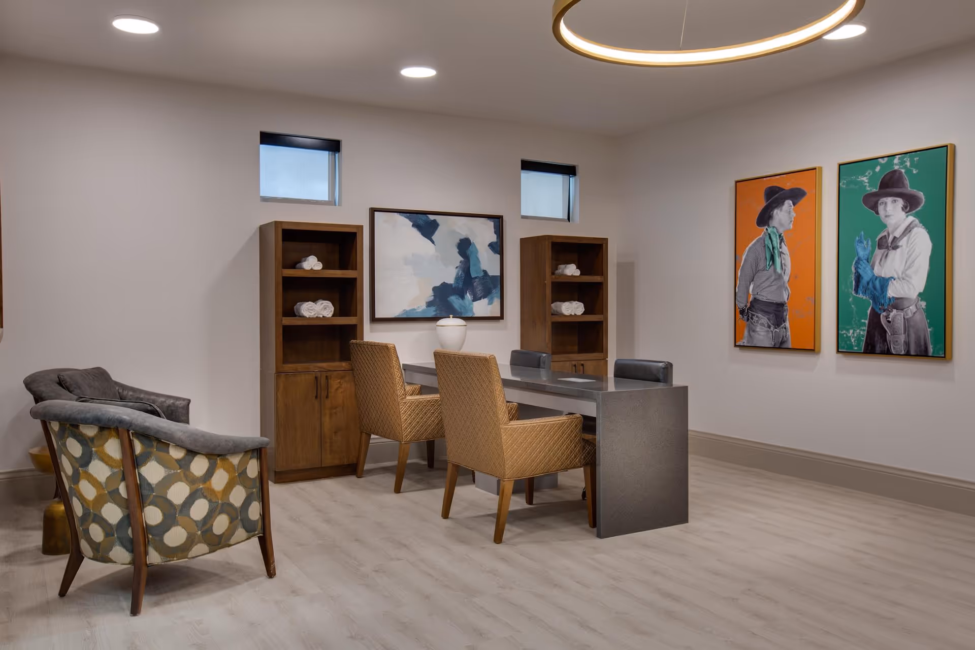A modern interior room with a sleek gray table surrounded by four chairs, two wooden shelves with rolled white towels, abstract and portrait artwork on the walls, and a patterned armchair in the foreground. The room has light wood flooring and recessed ceiling lights.