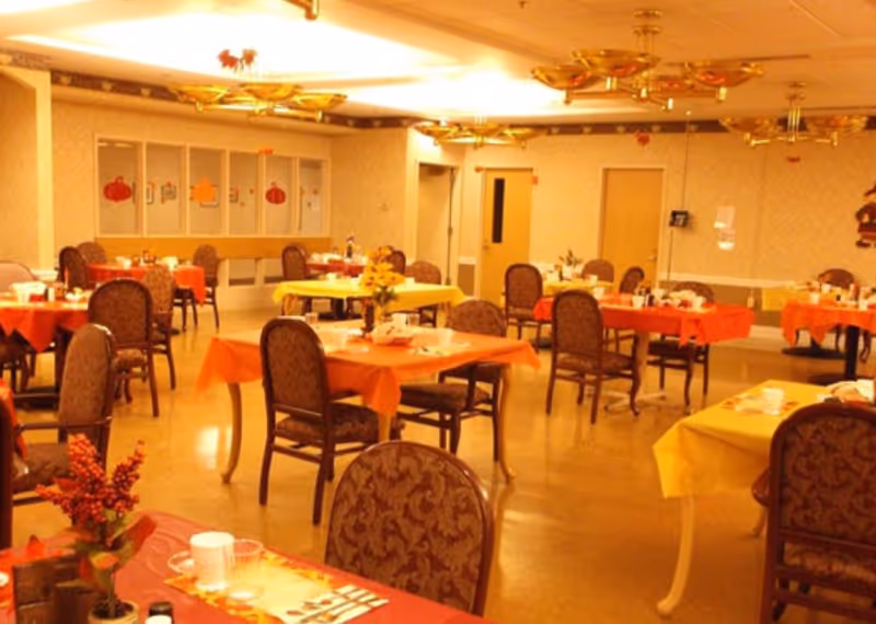 A dining room with multiple tables covered in orange and yellow tablecloths, each set with cups, plates, and utensils. The room has patterned chairs around the tables, warm lighting from ceiling fixtures, and autumn-themed decorations on the walls and tables.