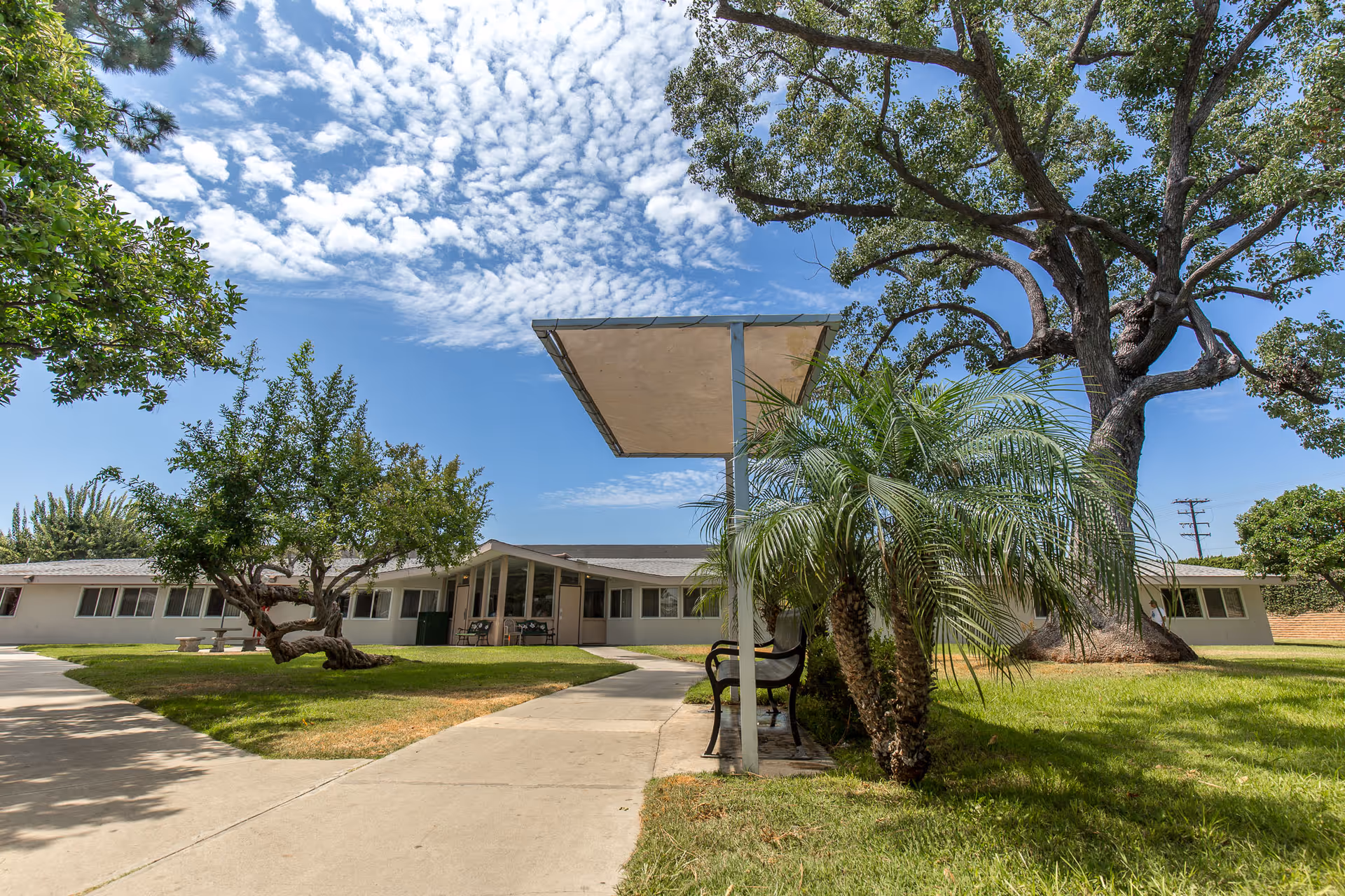 Front exterior of a single-story care facility with a walkway, covered bench, lawn and large trees under a blue sky.