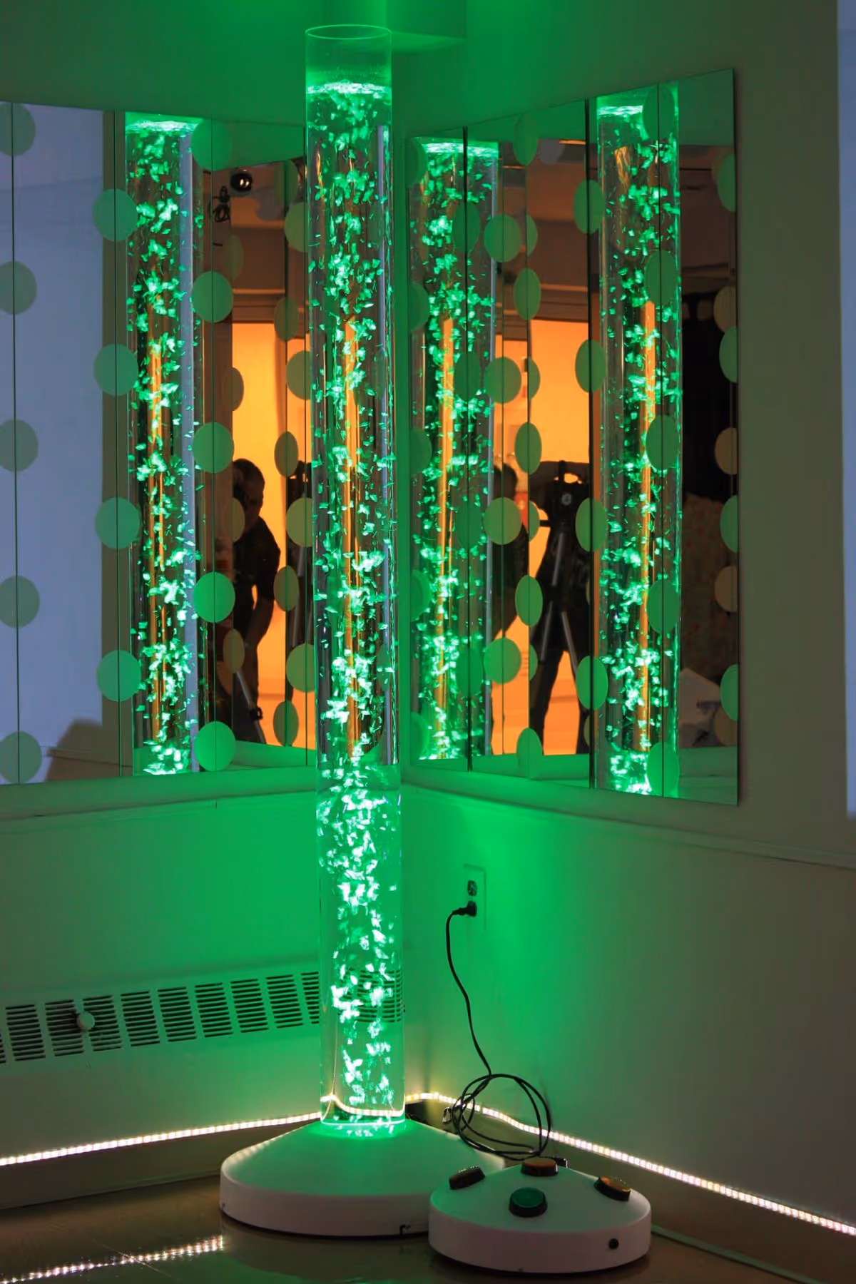 A tall cylindrical bubble tube lamp glowing green, placed in the corner of a room with mirrors on the walls reflecting the light and bubbles. The lamp is plugged into an electrical outlet and has a control base with buttons on the floor next to it.