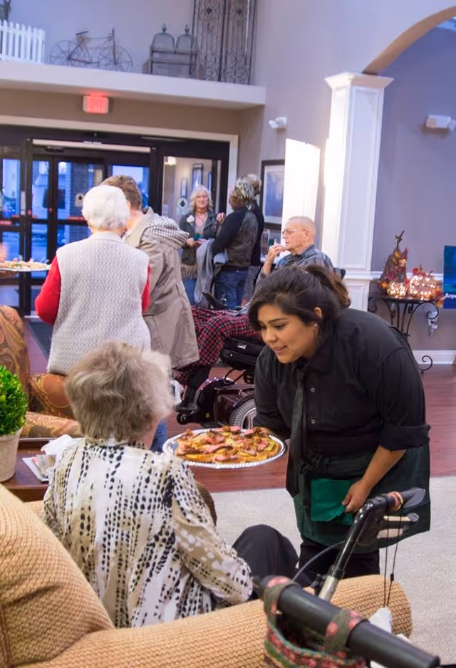 A caregiver in a black shirt and green apron is offering a plate of appetizers to an elderly woman seated on a beige couch in a senior living facility common area. Other elderly residents and visitors are seen socializing in the background near the entrance doors. The room has warm lighting, decorative plants, and comfortable seating.