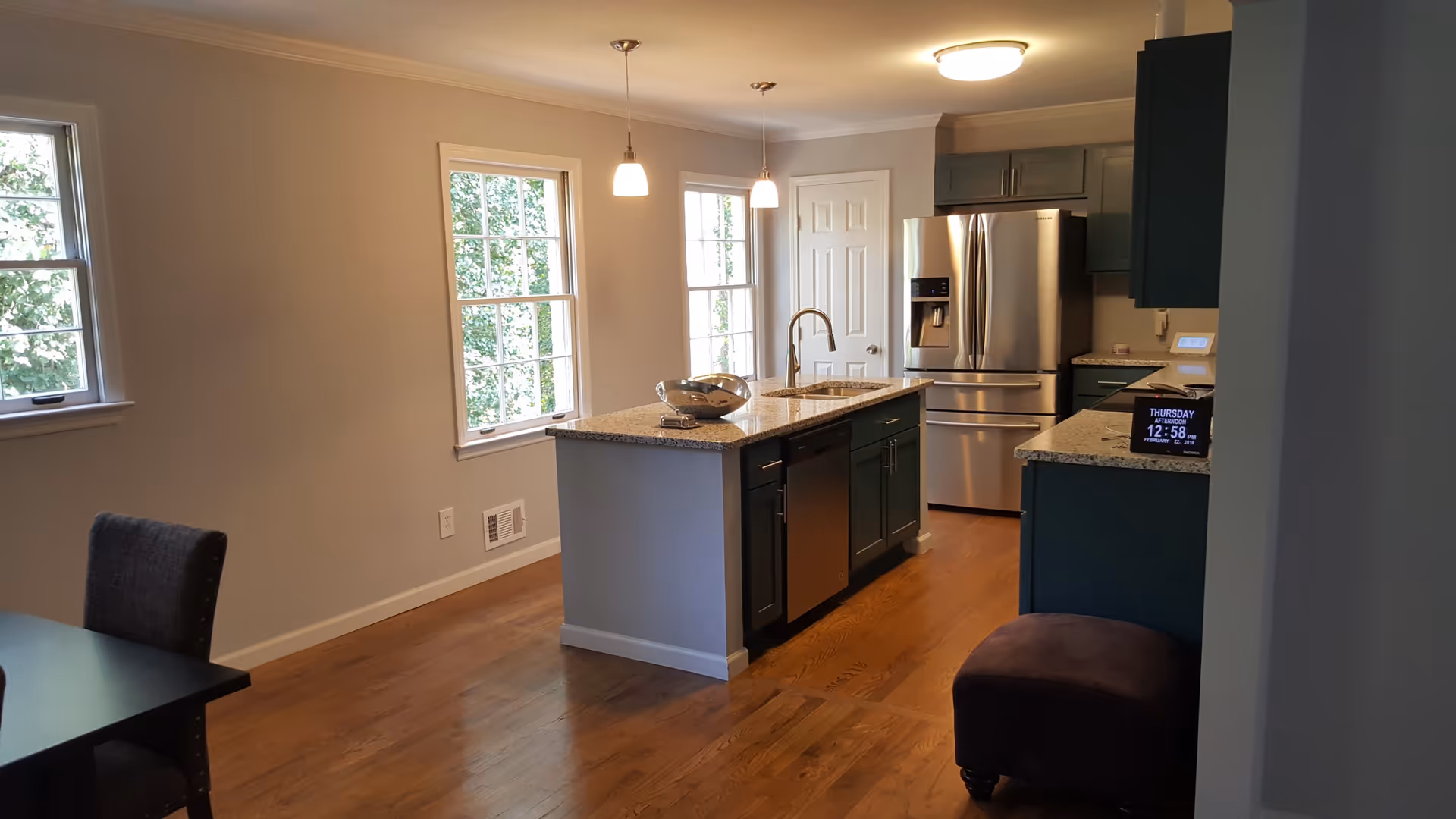 Modern kitchen with wooden floor, two pendant lights hanging over a granite countertop island with a sink and dishwasher. Stainless steel refrigerator and blue cabinets are visible. Two windows provide natural light, and a small digital clock is on the counter.