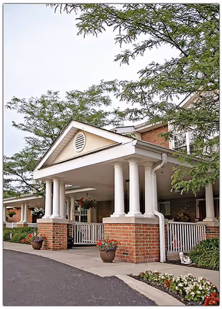 Front exterior view of a senior living facility with a covered entrance supported by white columns and brick pillars. There are potted plants and flowers near the entrance, and trees partially frame the building.
