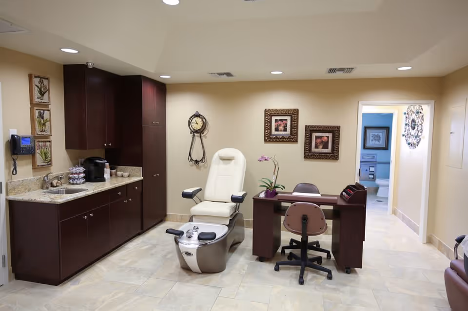 A well-lit room with beige walls and tiled floor featuring a pedicure spa chair with foot bath, a manicure table with two chairs, dark wood cabinets with a sink and coffee maker, decorative wall clock, and framed floral artwork. A doorway leads to a bathroom with a toilet visible.