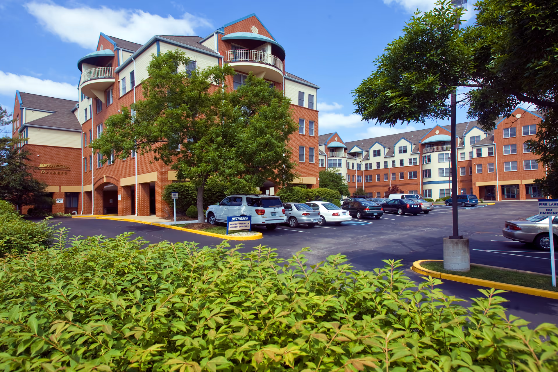 Front exterior of a multi-story brick senior living building with balconies, a parking lot with cars, and landscaped greenery.