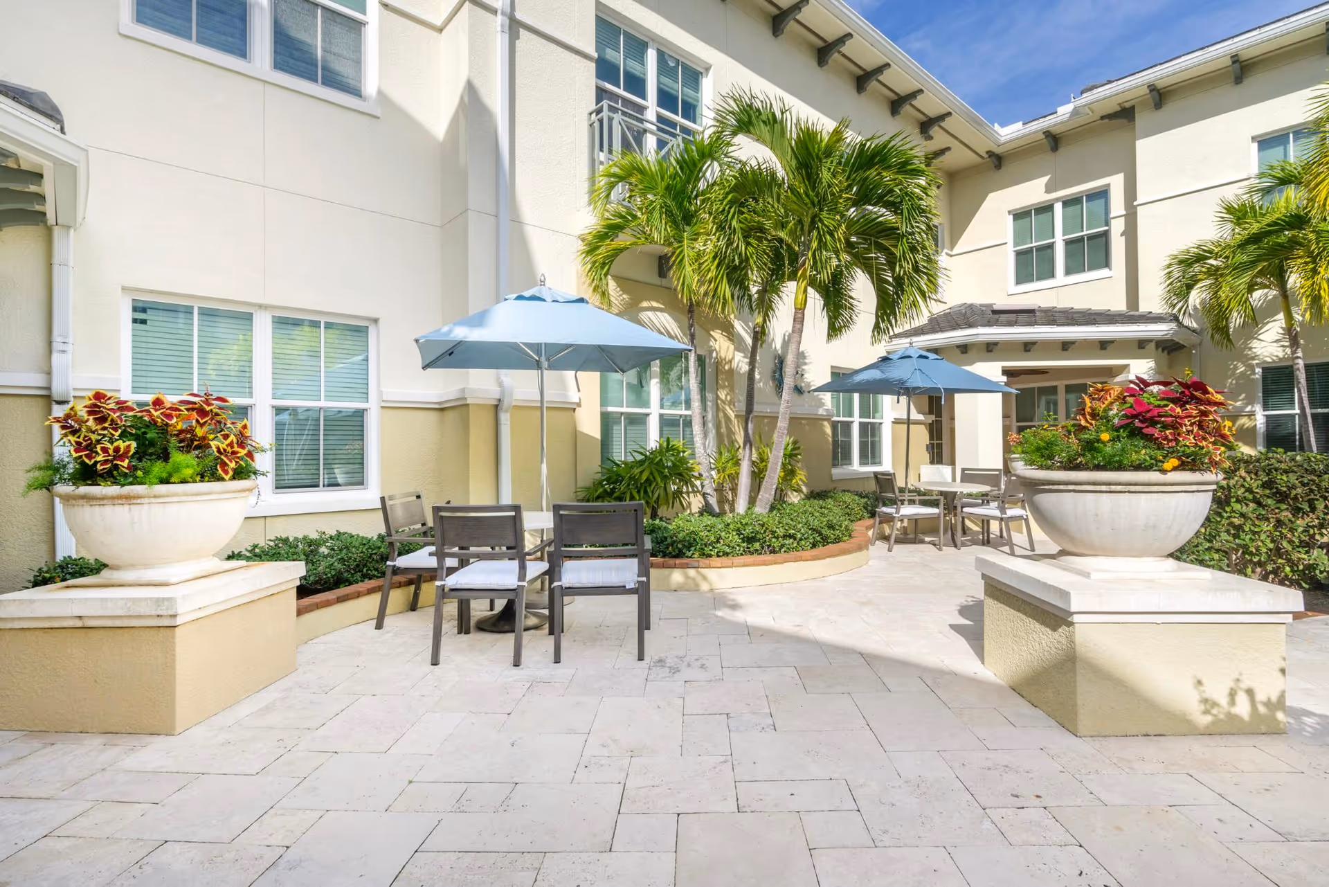 Outdoor patio area with beige tiled flooring, two large planters with colorful plants, several palm trees, and two sets of tables with chairs and blue umbrellas. The patio is surrounded by a light-colored building with multiple windows under a clear blue sky.