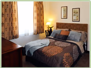 Cozy bedroom with a patterned bed, pillows, a bedside table and lamp next to a curtained window.