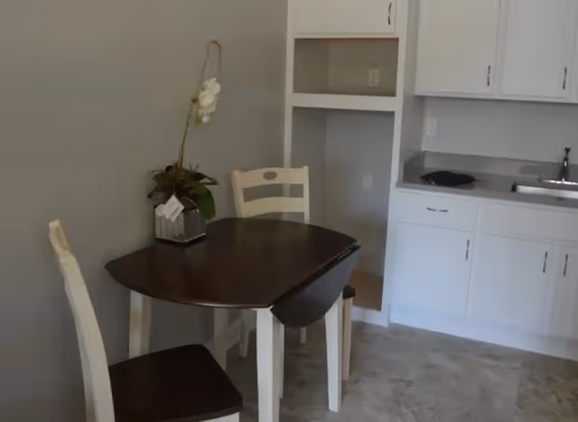 Small dining area with a dark wooden table and two white chairs with dark seats. A potted orchid plant is placed on the table. In the background, there is a kitchenette with white cabinets, a countertop, and a sink.