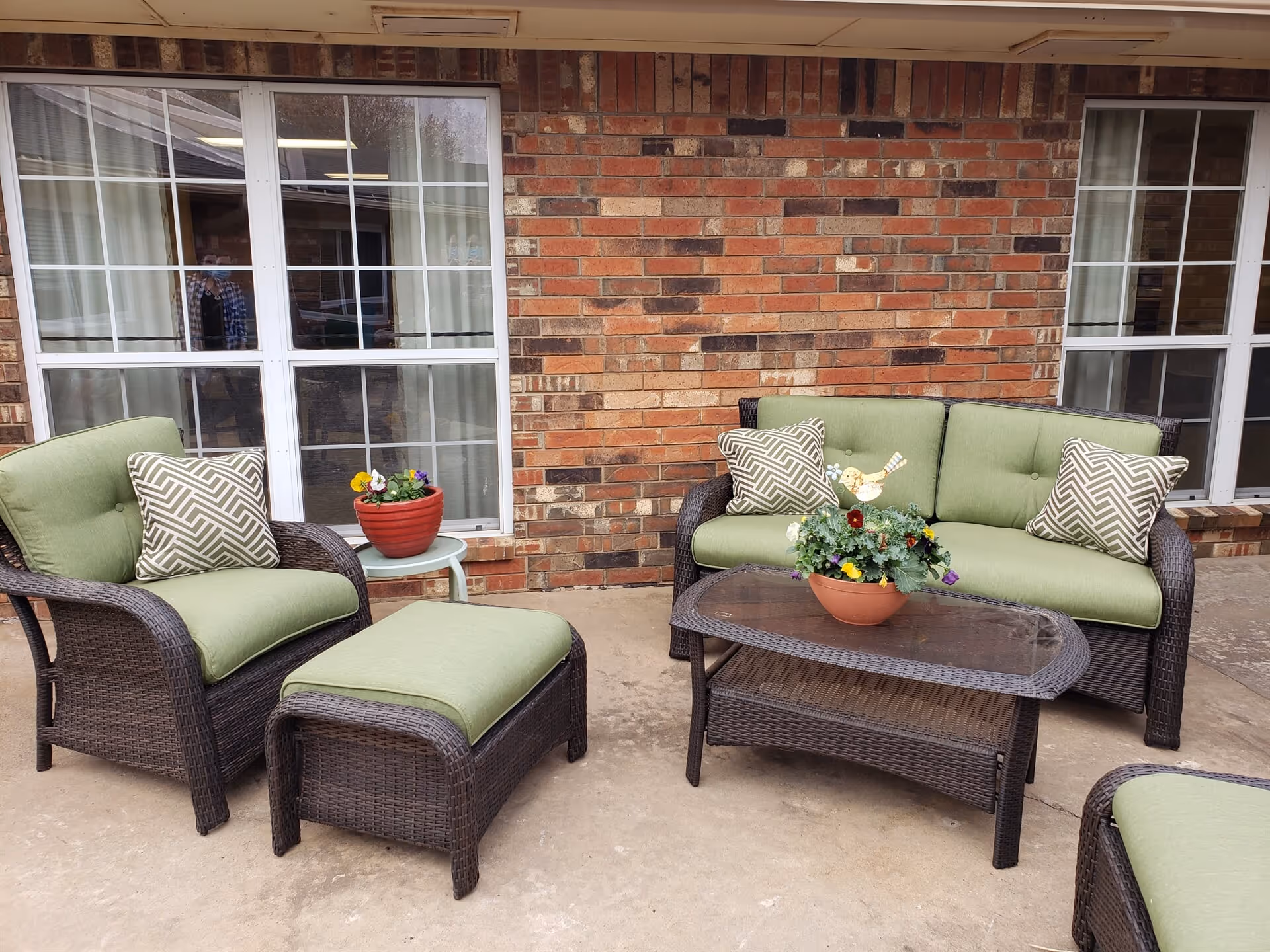 Outdoor patio area with brown wicker furniture including a cushioned armchair, ottoman, loveseat with green cushions and patterned pillows, and a glass-top coffee table with a flower pot. The background shows a brick wall with two large windows.
