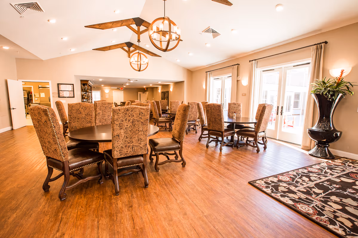 A spacious dining room with multiple round tables surrounded by upholstered chairs with wooden legs. The room has wooden flooring, beige walls, large windows with curtains, and two spherical chandeliers hanging from a vaulted ceiling with wooden beams. A large decorative vase with plants is placed near the windows.