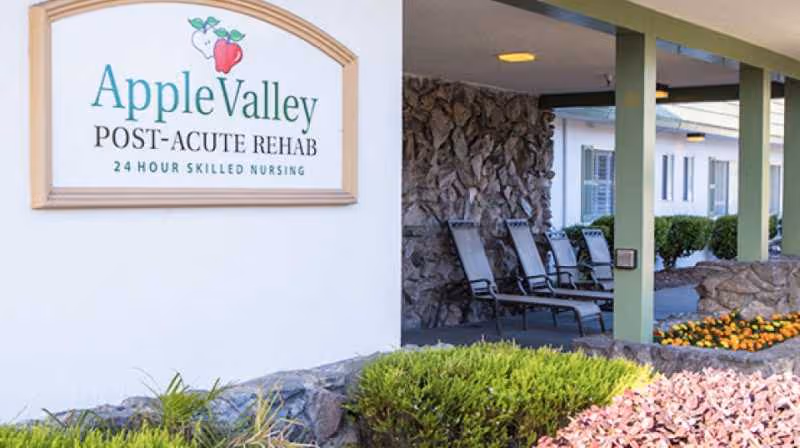 Exterior of Apple Valley Post-Acute Rehab with sign, covered walkway, and empty chairs.