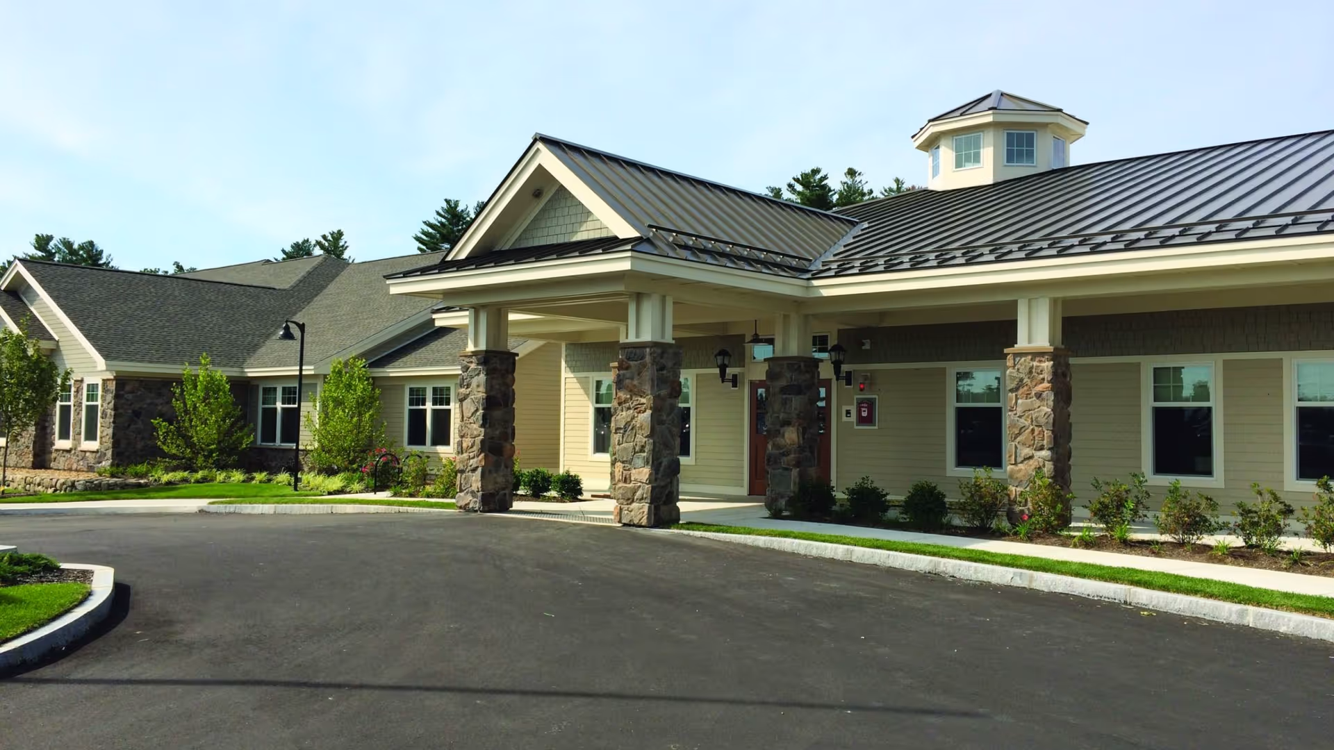 Exterior view of a senior living facility building with stone pillars supporting a covered entrance. The building has beige siding, multiple windows, and a metal roof with a small cupola on top. There is a paved driveway and landscaped greenery around the building.