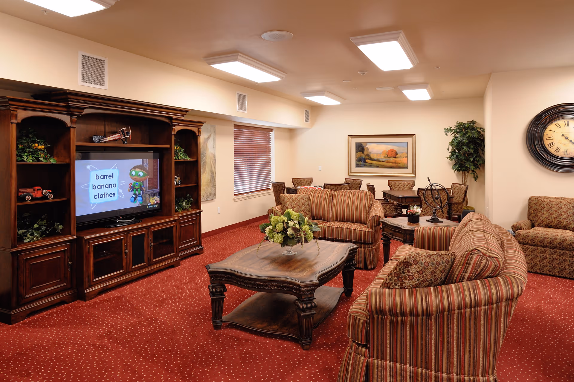 A cozy living room area in a senior living facility with striped upholstered sofas and armchairs arranged around a wooden coffee table with a floral centerpiece. A large wooden entertainment center holds a television displaying a children's educational program. The room has red carpet, beige walls, a large wall clock, framed artwork, and a potted plant in the corner.
