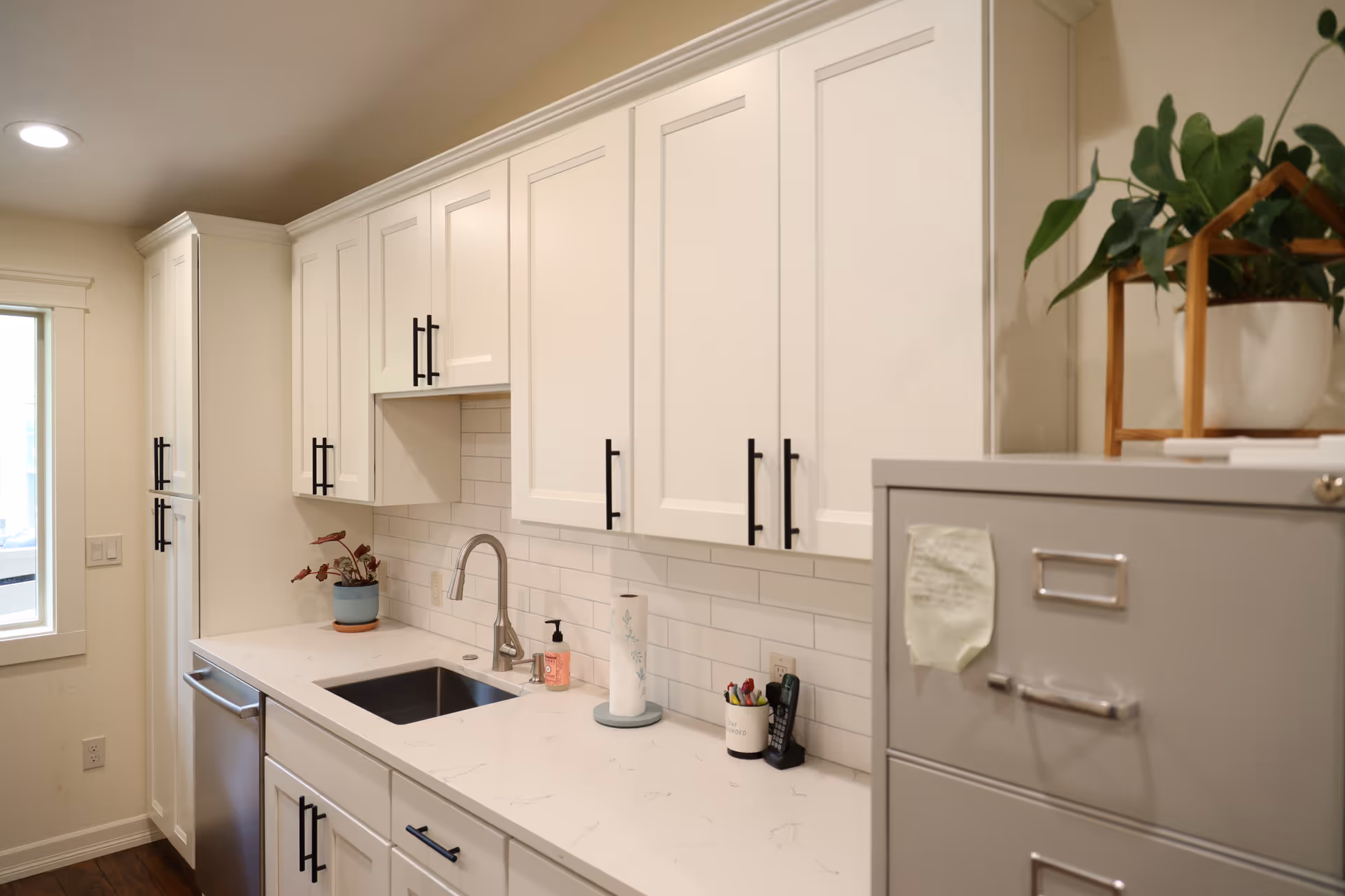 A modern kitchen with white cabinets and black handles, a stainless steel sink with a faucet, a dishwasher, a white countertop with a paper towel holder, hand soap, a small potted plant, and a gray filing cabinet with a potted plant on top.