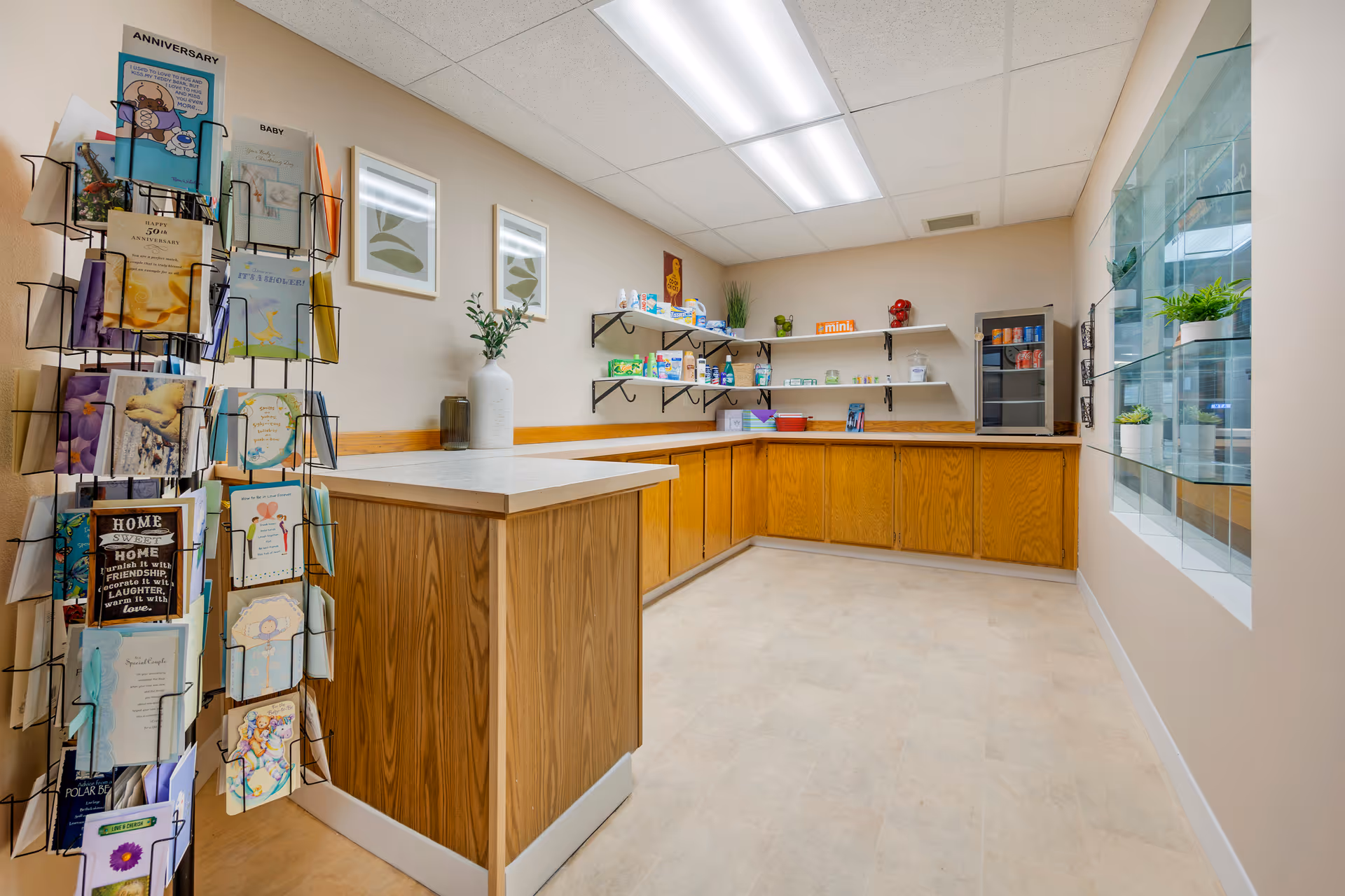Interior view of a small snack and greeting card area in a senior living facility. The room features wooden cabinets with a countertop, open shelves stocked with snacks and small items, a small refrigerator with drinks, and a rotating rack filled with various greeting cards. The walls are beige, and there are two framed leaf prints hanging on one wall. A glass display case with small potted plants is visible on the right side.