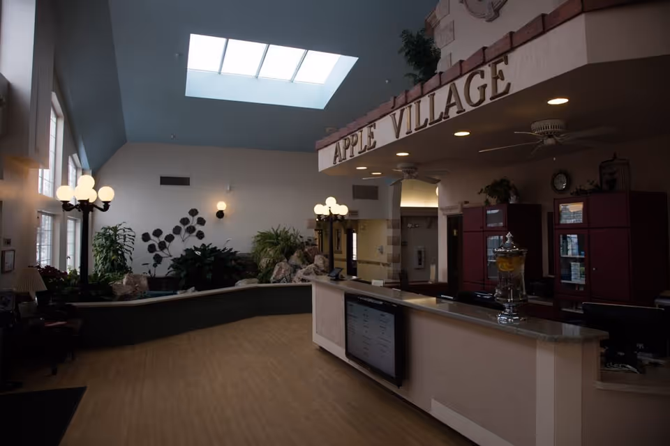 Lobby reception area with a front desk, indoor plants, skylight, and a large 'Apple Village' sign overhead.