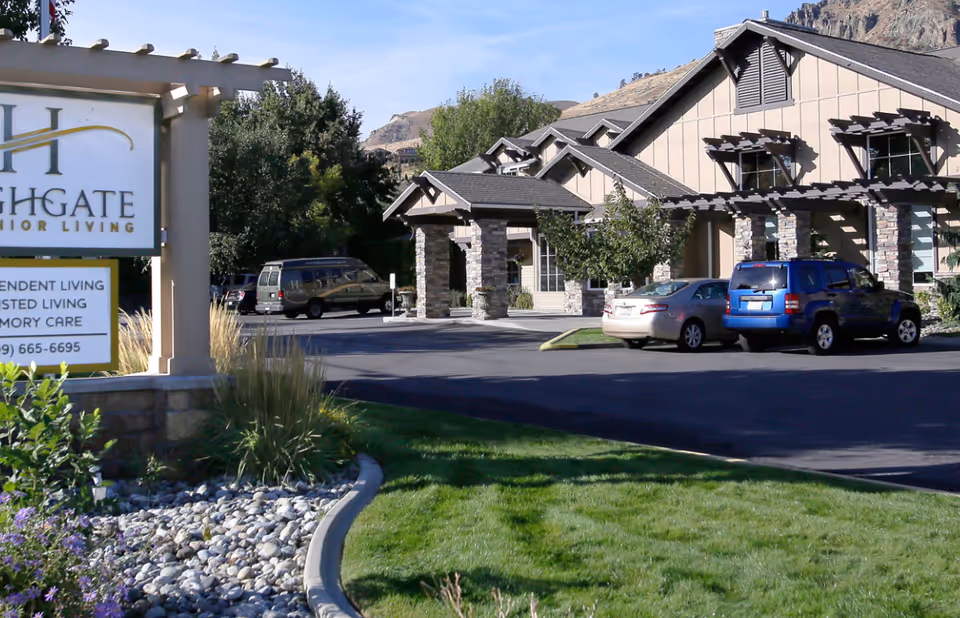 Exterior view of Highgate Senior Living facility showing the building with stone pillars and a parking lot with cars. There is a sign in the foreground with the facility name and contact information, surrounded by landscaping with grass, rocks, and plants. Hills and trees are visible in the background under a clear blue sky.