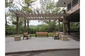 Outdoor seating area with a wooden pergola and three benches overlooking a lawn beside a building.