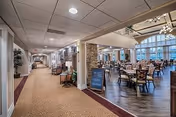 Interior view of a senior living facility showing a wide hallway with carpet flooring on the left and a dining area with wooden tables and chairs on the right. The dining area has large windows allowing natural light to fill the space. There is a small chalkboard sign near the entrance to the dining area and decorative plants along the hallway.