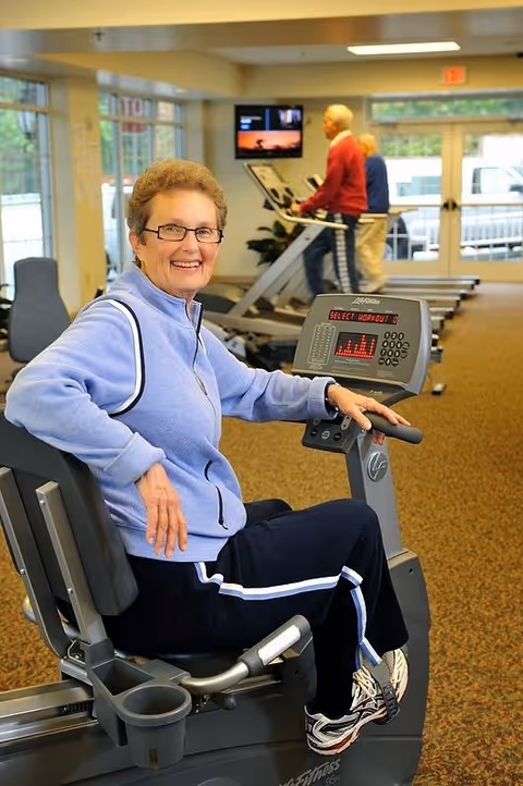 An elderly woman wearing glasses and a light blue jacket is sitting on a stationary exercise bike in a fitness room. She is smiling at the camera. In the background, two elderly men are using treadmills near large windows and glass doors.