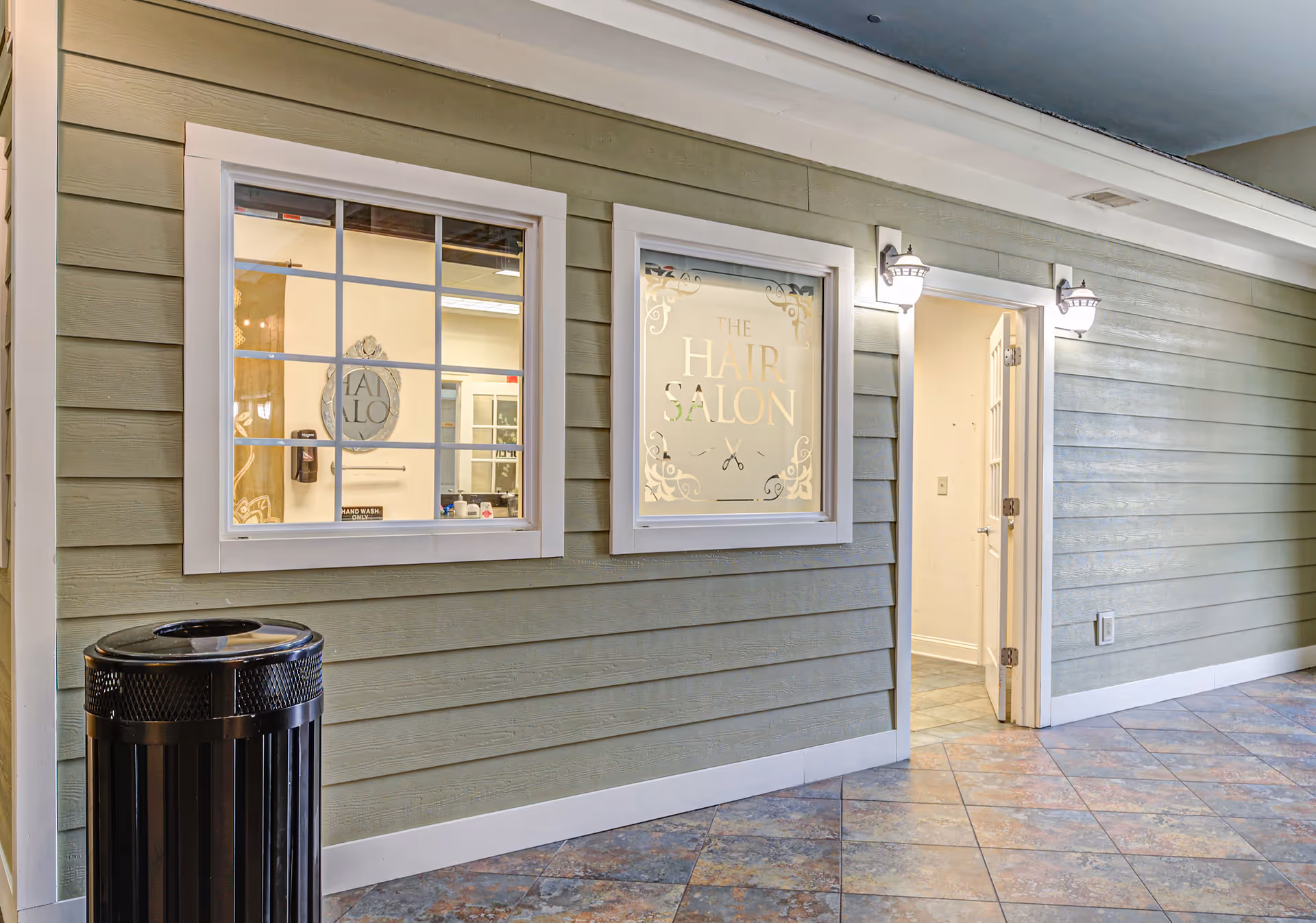 Interior hallway with green horizontal siding walls, two white-framed windows, and an open door leading to a room. One window has frosted glass with decorative text reading 'The Hair Salon' and a scissors icon. The other window shows a mirror and some salon items inside. Two wall-mounted lights are above the door and window. A black trash can is positioned near the corner.