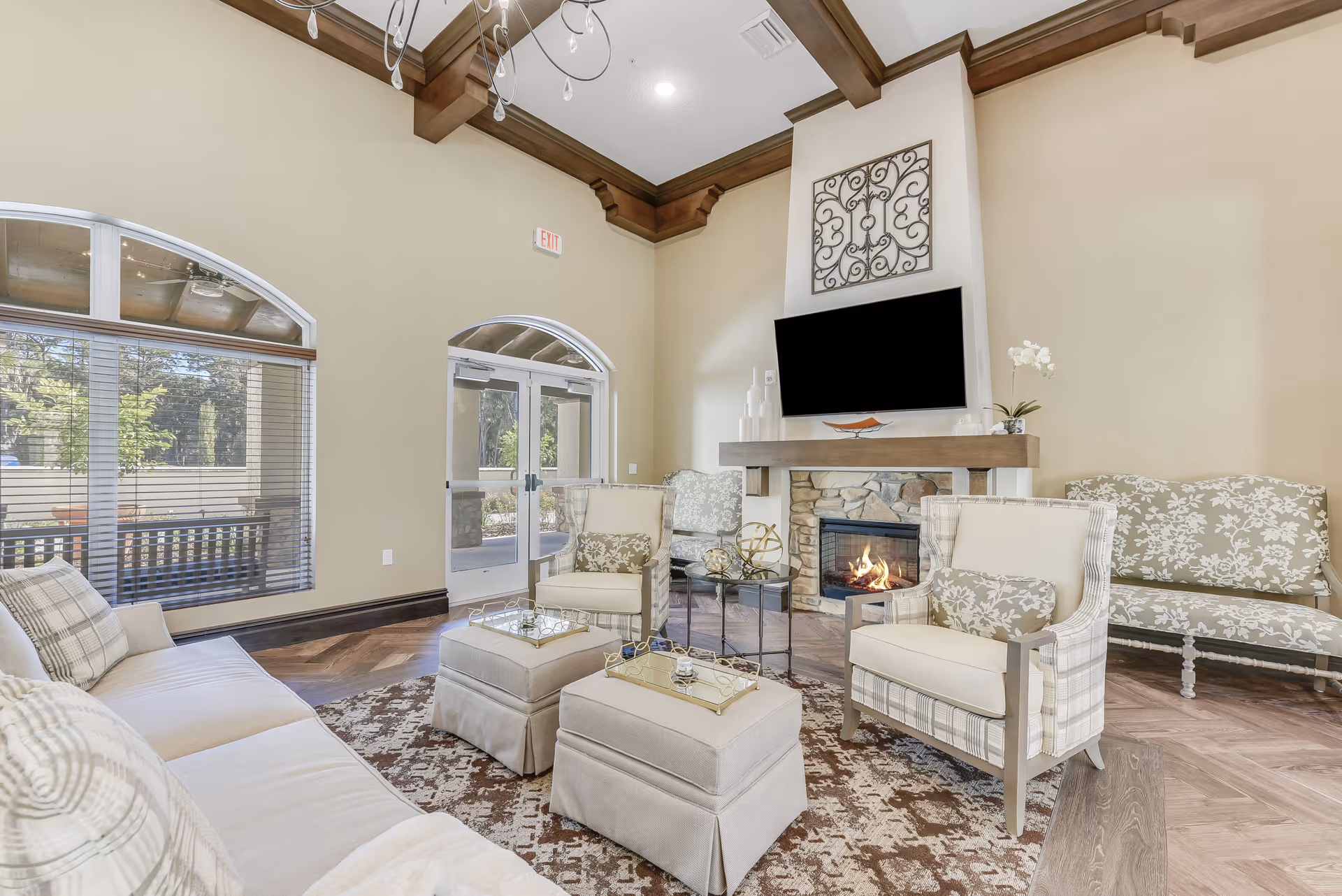 A cozy senior living facility common area with a high ceiling and wooden crown molding. The room features a stone fireplace with a mounted flat-screen TV above it, surrounded by comfortable upholstered chairs and a sofa in neutral tones. There are two ottomans in the center on a patterned rug, a glass coffee table with decorative items, large windows with blinds letting in natural light, and a glass door leading outside.