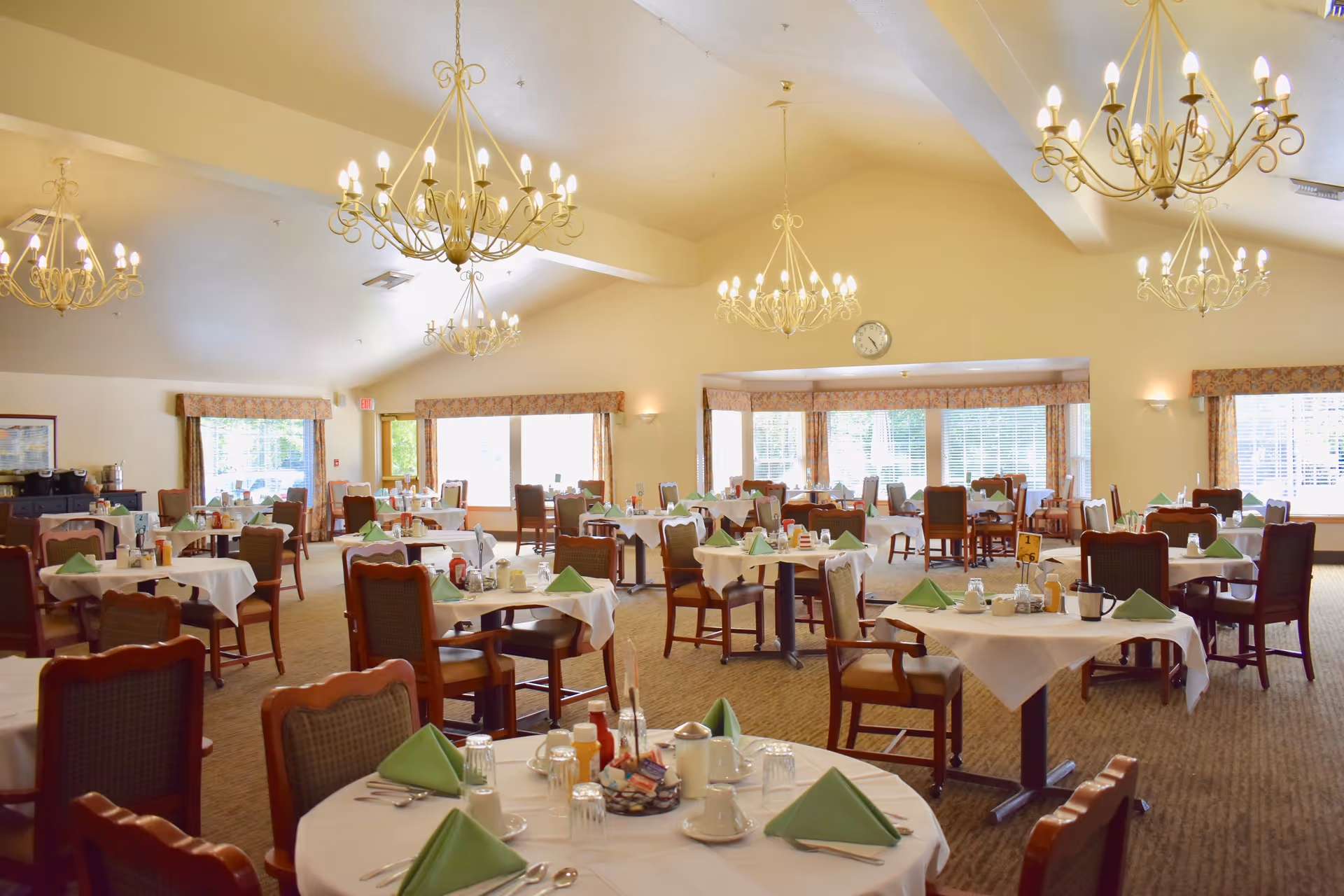 A spacious dining room with multiple round tables covered with white tablecloths, each set with green folded napkins, cups, glasses, and condiments. The room features large windows with curtains, beige walls, carpeted floors, and several elegant chandeliers hanging from the ceiling.