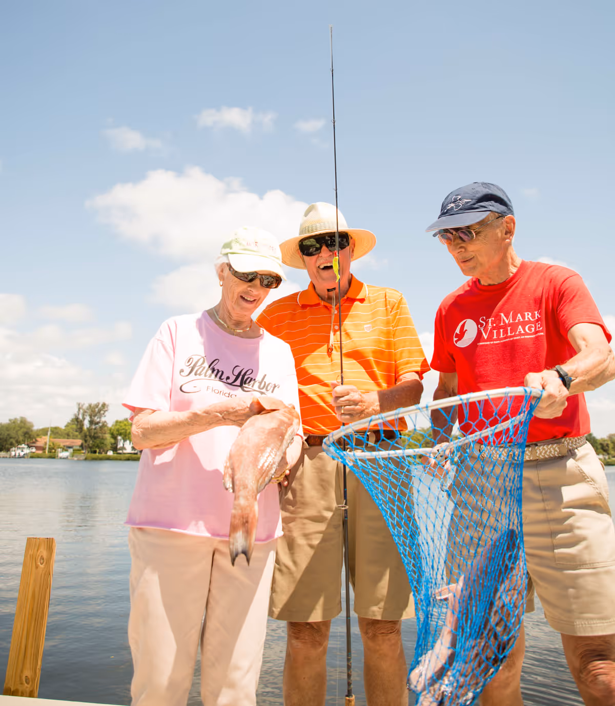 Three elderly people standing on a dock by a lake on a sunny day. One woman in a pink Palm Harbor t-shirt and light pants is holding a fish. A man in an orange striped polo shirt and beige shorts is holding a fishing rod. Another man in a red St. Mark Village t-shirt and beige shorts is holding a blue fishing net with a fish inside. They are all wearing hats and sunglasses and appear to be enjoying fishing together.