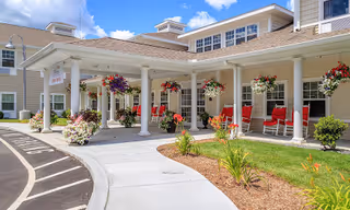 Front exterior view of All American Assisted Living at Warwick showing a covered entrance with white columns, hanging flower baskets, red rocking chairs, and landscaped garden beds under a blue sky with some clouds.