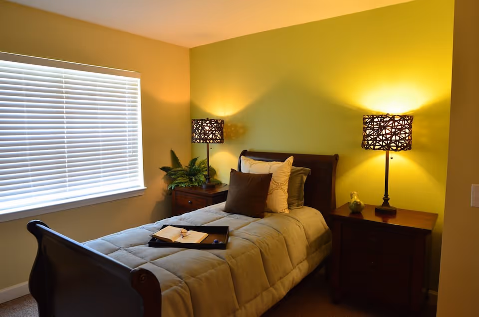 A cozy bedroom with a single wooden bed featuring a gray comforter and multiple pillows. There are two matching wooden nightstands on either side of the bed, each with a decorative lamp emitting warm light. A window with closed white blinds is on the left wall, and a small plant is placed on the left nightstand.