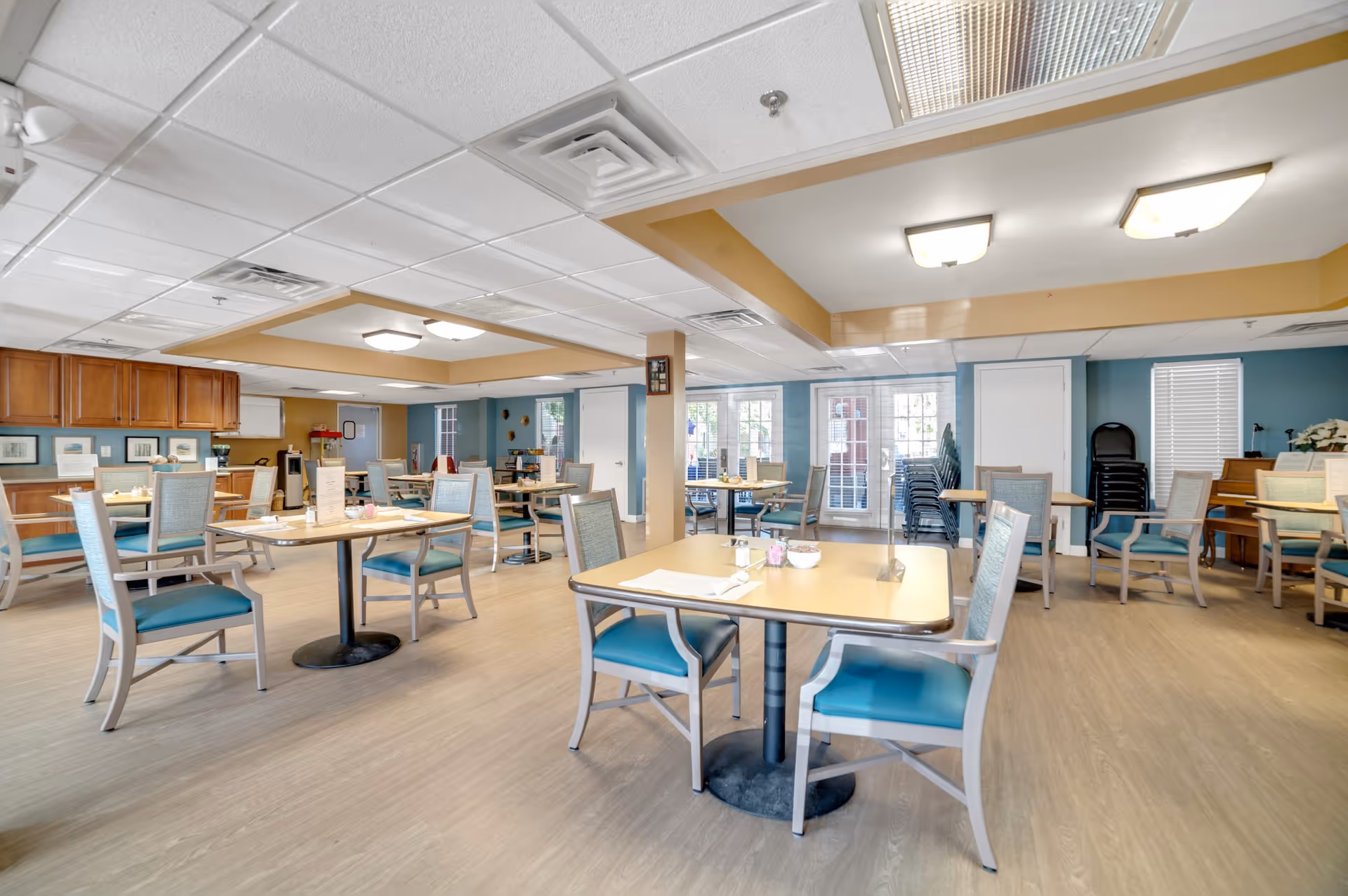 Bright communal dining room with multiple tables and teal-upholstered chairs, wood-look flooring, and a service counter in the back.