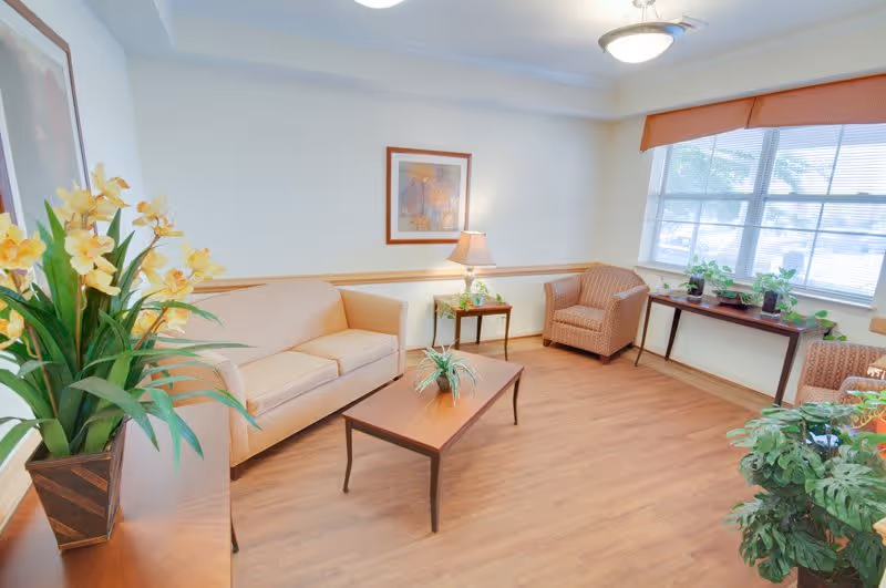 A bright and cozy living room with a beige sofa, a matching armchair, a wooden coffee table with a small plant, and a side table with a lamp. There are framed pictures on the walls, large windows with blinds letting in natural light, and several potted plants placed around the room.
