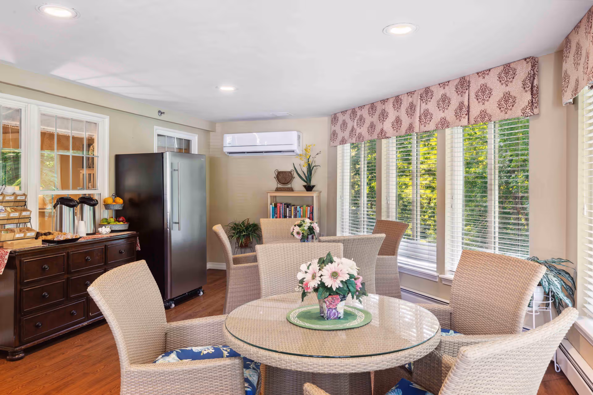 A bright and cozy dining area with a round glass-top table surrounded by wicker chairs with blue floral cushions. The room features large windows with white blinds and patterned valances, allowing natural light to fill the space. A dark wooden sideboard holds coffee dispensers, fruit, and snacks. A stainless steel refrigerator stands against the wall, next to a small bookshelf with plants and decorative items. The floor is wooden, and the walls are painted a soft beige.