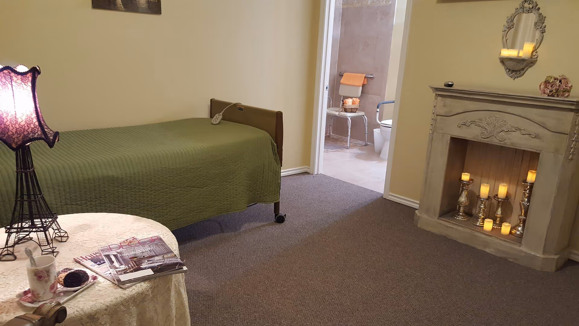 A cozy senior living bedroom with a single bed covered in a green quilt, a small round table with a lace tablecloth holding a cup, a pastry, and magazines. A decorative lamp with a purple shade is on the table. There is a faux fireplace with multiple lit candles and a mirror above it. An open door reveals a bathroom with a toilet, a chair, and a towel rack with an orange towel.