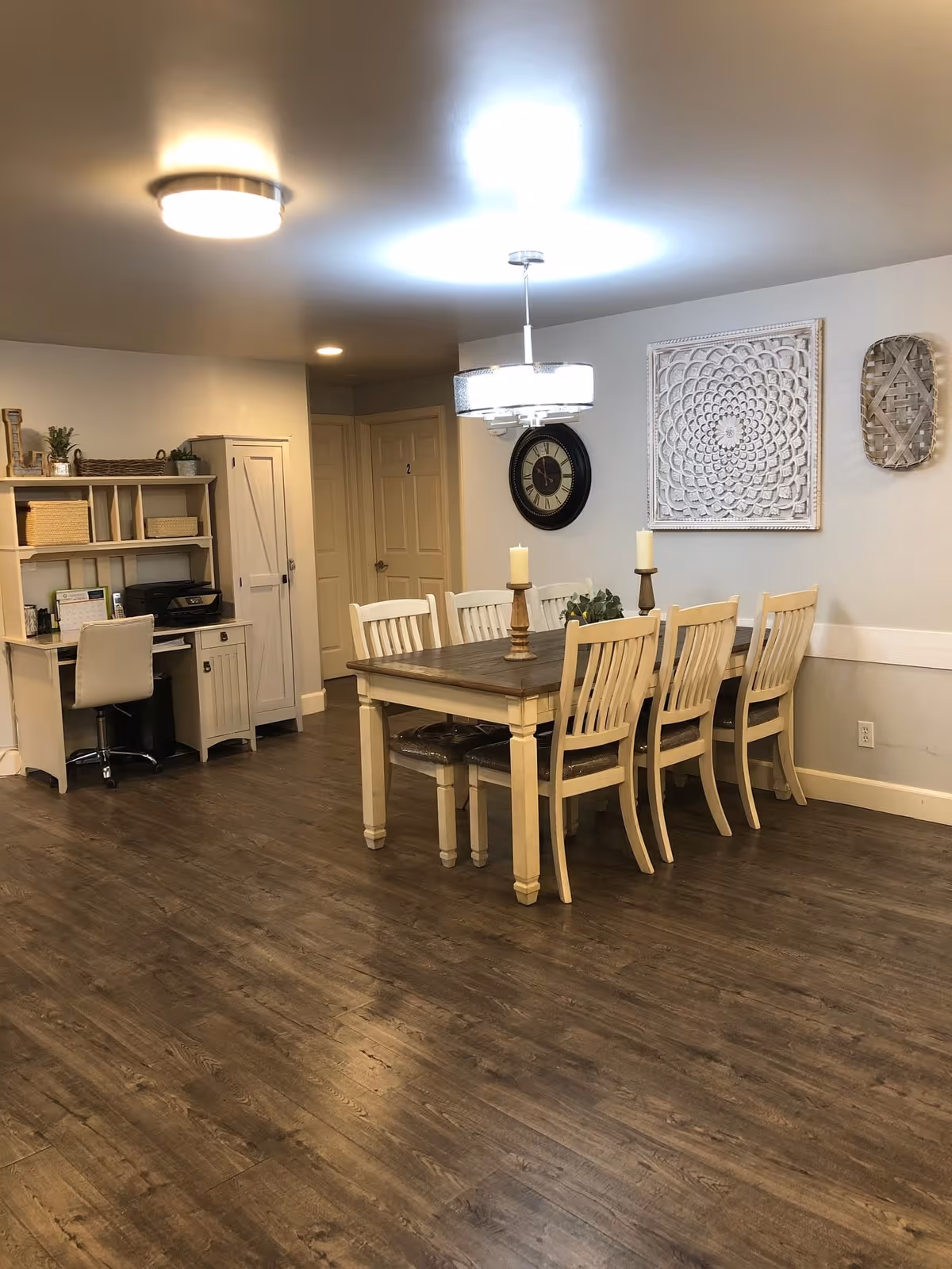 A dining area with a wooden table and six matching chairs, two candles on the table, a wall clock, and decorative wall art. To the left, there is a white desk with a chair, shelves, and a printer. The floor is wooden, and the walls are light-colored.