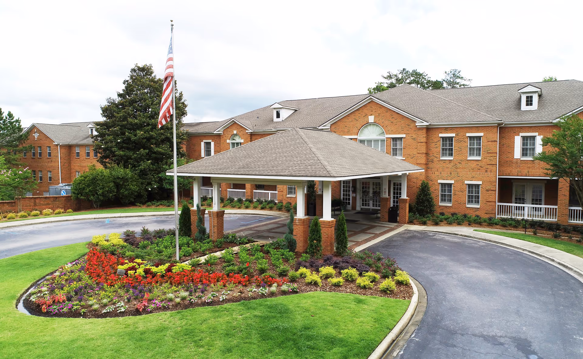Front exterior view of a large brick senior living facility with a covered entrance driveway, a landscaped flower bed with colorful flowers, and an American flag on a flagpole.