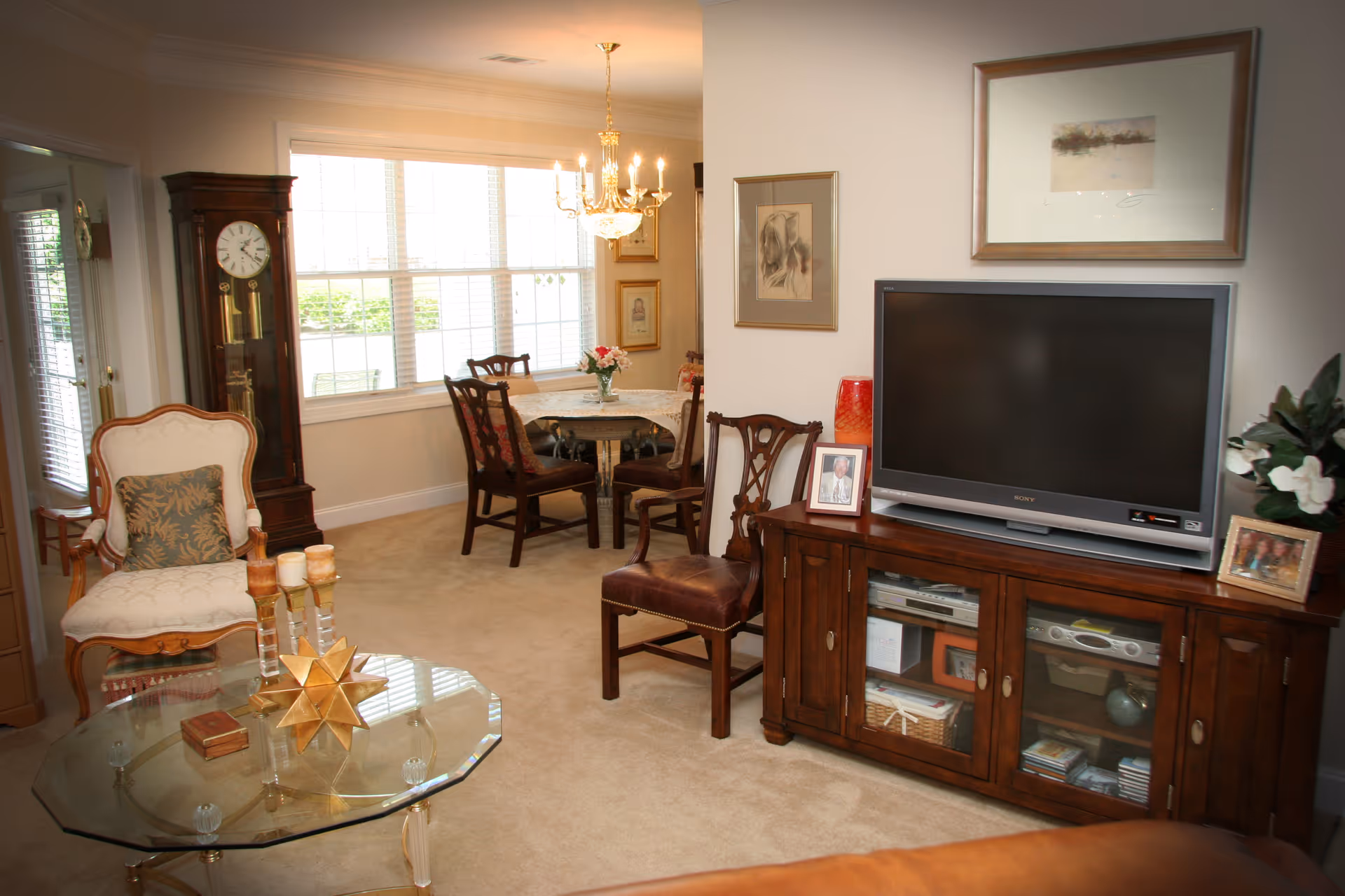 A cozy living room and dining area with beige carpet and walls. The living room features a glass coffee table with decorative items, an upholstered armchair with a patterned pillow, a wooden grandfather clock, and a wooden TV stand with a flat-screen television and framed photos. The dining area has a round table with four wooden chairs, a vase with flowers, and a chandelier hanging above. Large windows allow natural light to fill the space.