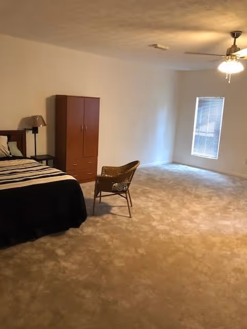 A spacious bedroom with beige carpet, a bed with striped bedding on the left, a wooden wardrobe, a small side table with a lamp, a wicker chair in the center, a window with blinds on the right wall, and a ceiling fan with lights.