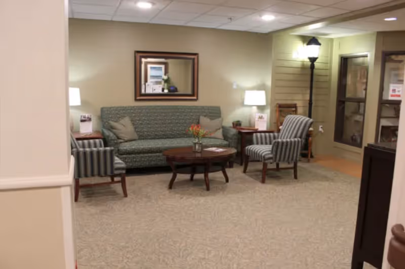 A cozy sitting area in a senior living facility featuring a green patterned sofa with two beige pillows, two striped armchairs, a wooden coffee table with a small flower arrangement, two side tables each with a lamp, a framed mirror on the wall, and a tall floor lamp near a glass door.