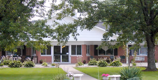 Front exterior view of a single-story brick building with white trim, surrounded by green grass, trees, and shrubs. A concrete walkway leads to the entrance with glass doors and windows on either side.